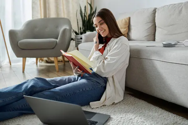 A young woman sits on the floor, working on her laptop, talking on the phone, and reading a notebook. — Stock Photo