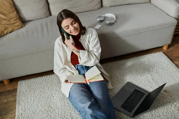 A young woman in casual attire sits on a carpet in front of a couch, working on her laptop while on a phone call. — Stock Photo