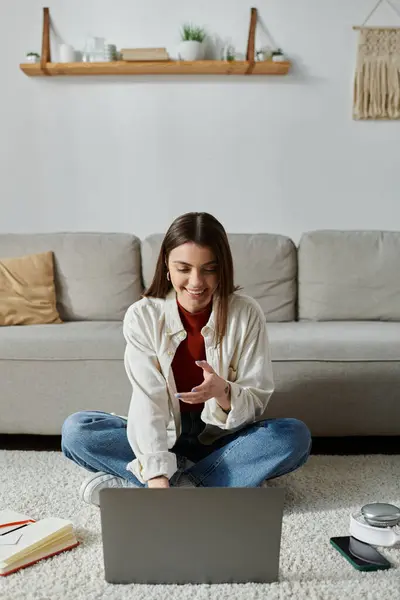 A young woman in casual attire sits on the floor, working on her laptop while gesturing with her hand. — Stock Photo