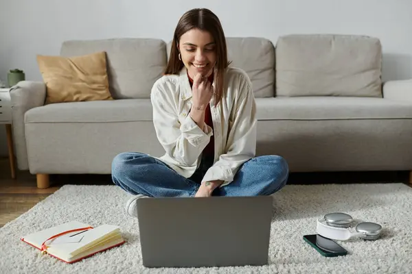 Una mujer joven se sienta en una alfombra esponjosa frente a un sofá, sonriendo mientras trabaja en su computadora portátil. - foto de stock