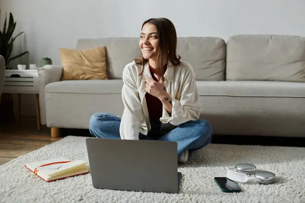 A young woman in casual attire sits on the floor in front of a couch, working on her laptop with a confident smile. — Stock Photo