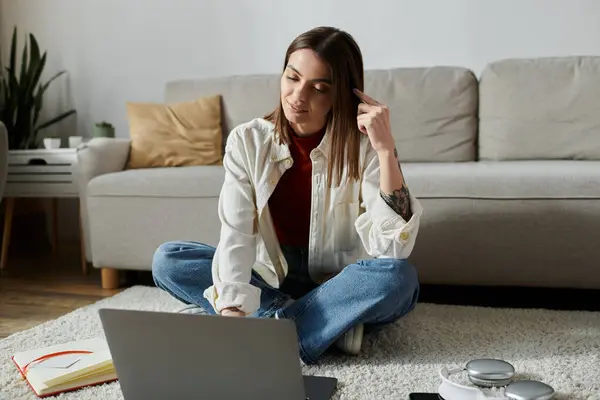 A young woman sits on a rug in front of a sofa, working on a laptop and looking thoughtful. — Stock Photo