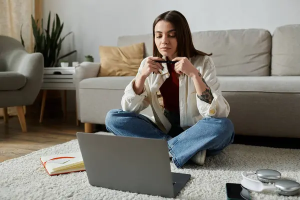 Eine junge Frau in Freizeitkleidung sitzt im Schneidersitz auf einem Teppich in ihrem Haus und arbeitet an ihrem Laptop. — Stockfoto