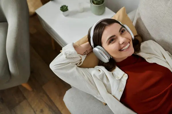 Una joven con los auriculares puestos, vistiendo una camisa roja y blanca, sonríe mientras se recuesta en un sofá. - foto de stock