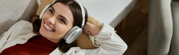 A young woman with headphones on, smiles while resting on her couch. — Stock Photo