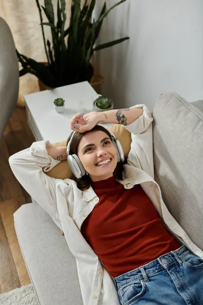 A young woman in casual attire relaxes on a couch with headphones on smiling at home — Stock Photo