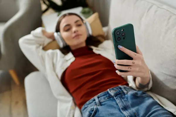 A young woman lies on a couch, using her phone, while wearing headphones. — Stock Photo