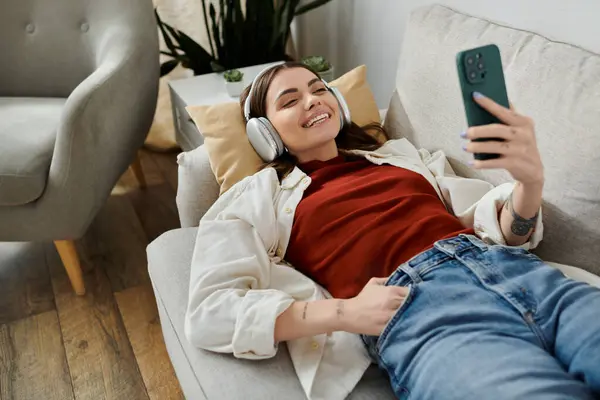 A young woman in casual clothes relaxes on a couch while looking at her phone and smiling. — Stock Photo