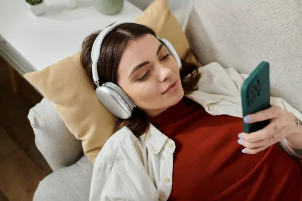 Uma jovem mulher de roupas casuais trabalha remotamente em casa, usando fones de ouvido e usando seu smartphone. — Fotografia de Stock