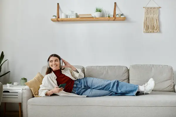 Uma jovem mulher de traje casual jaz em um sofá, fones de ouvido, segurando um smartphone. Ela está trabalhando remotamente de casa. — Fotografia de Stock