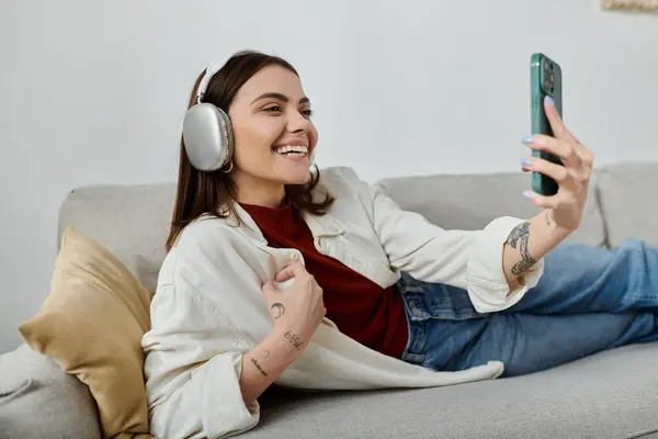 Uma jovem mulher em traje casual relaxa em um sofá enquanto trabalha remotamente em seu telefone, sorrindo brilhantemente. — Fotografia de Stock