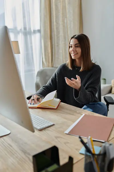 A young woman in a grey sweater sits at her desk, engaged in a video call while reviewing a notebook. — Stock Photo