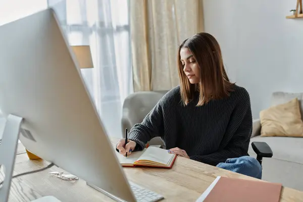A young woman in a grey sweater sits at a desk in her home office, working on a content strategy while writing in a notebook. — Stock Photo
