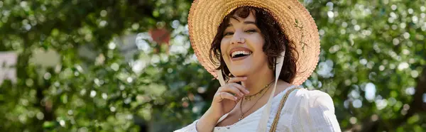 A brunette woman smiles brightly, wearing a straw hat and white blouse in a summer garden. — Stock Photo