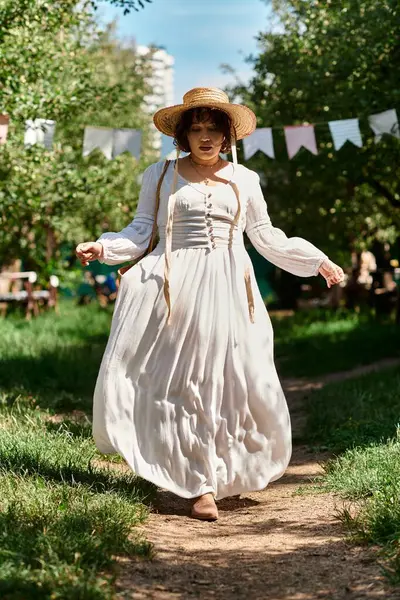 A young woman in a white dress and straw hat walks along a path in a summer garden. — Stock Photo