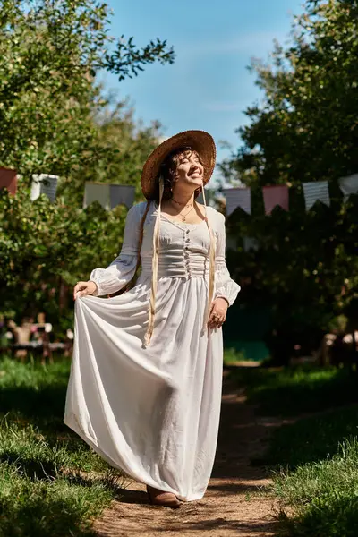 A young woman in a white dress and straw hat walks along a path in a garden, surrounded by green trees and blue sky. — Stock Photo