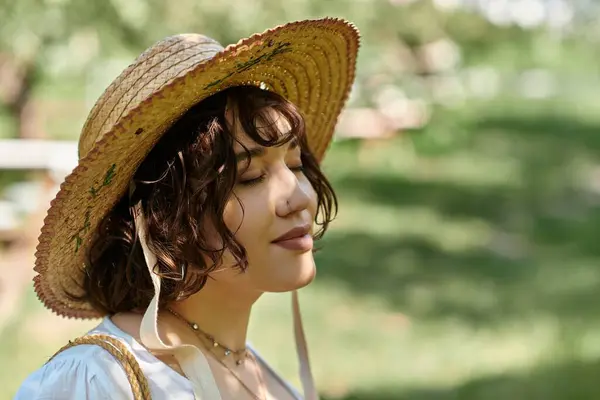A young woman with brunette hair in a straw hat and white blouse relaxes in a sunny garden, eyes closed in peaceful contemplation. — Stock Photo