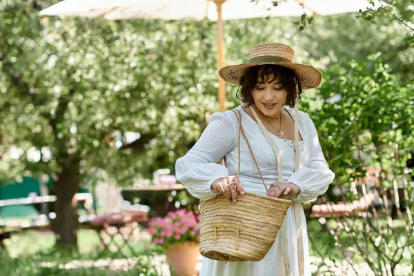 Una donna bruna con un cappello di paglia e una camicetta bianca passeggia in un giardino estivo, tenendo in mano un cestino tessuto. — Foto stock