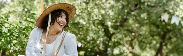 Une jeune femme en chemisier blanc et chapeau de paille sourit joyeusement en marchant dans un jardin d'été luxuriant. — Photo de stock