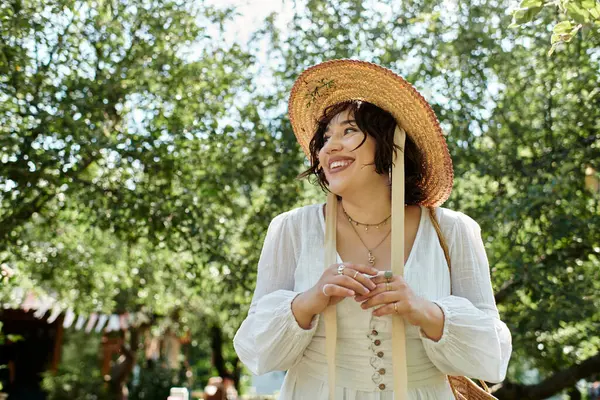 Eine brünette Frau mit Strohhut und weißer Bluse lächelt strahlend in einem Sommergarten. — Stockfoto