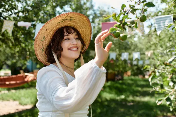 A young woman with dark hair, wearing a straw hat and a white blouse, reaches up to pluck fruit from a tree in a lush summer garden. — Stock Photo