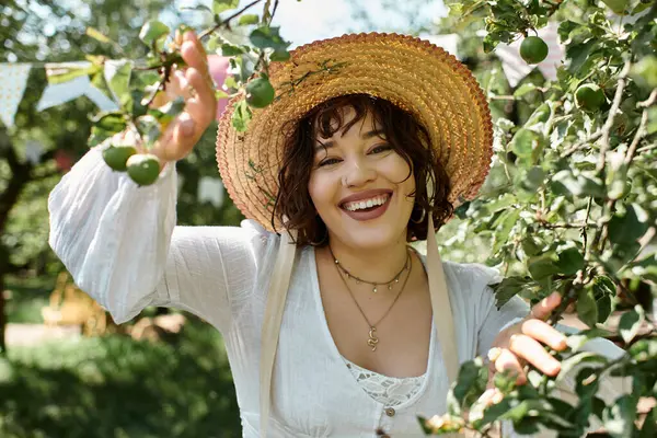 Uma jovem mulher de blusa branca e chapéu de palha sorri brilhantemente enquanto desfruta da beleza de um jardim de verão. — Fotografia de Stock