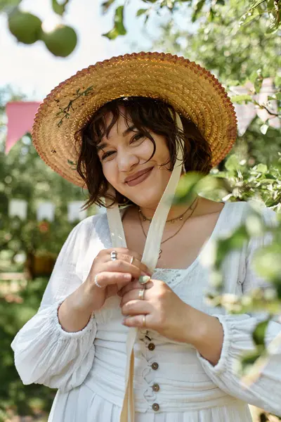 A young woman in a white dress and straw hat smiles softly in a garden setting. — Stock Photo
