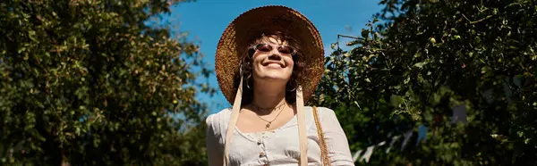 A young woman wearing a straw hat and sunglasses smiles brightly as she walks through a summer garden. — Stock Photo