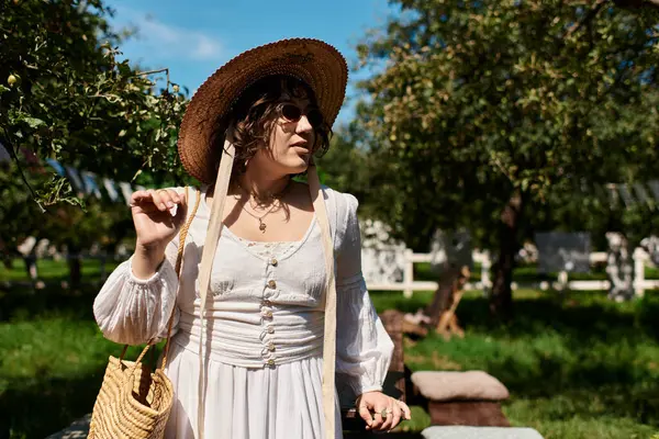 Брюнетка в солом'яному капелюсі і біла сукня прогулюється крізь пишний літній сад. — стокове фото