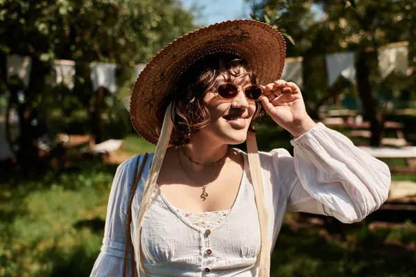 A brunette woman in a straw hat and white blouse smiles while adjusting her sunglasses in a summer garden. — Stock Photo