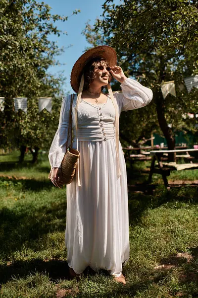 A brunette woman in a white dress and straw hat stands in a lush summer garden, bathed in sunlight. — Stock Photo