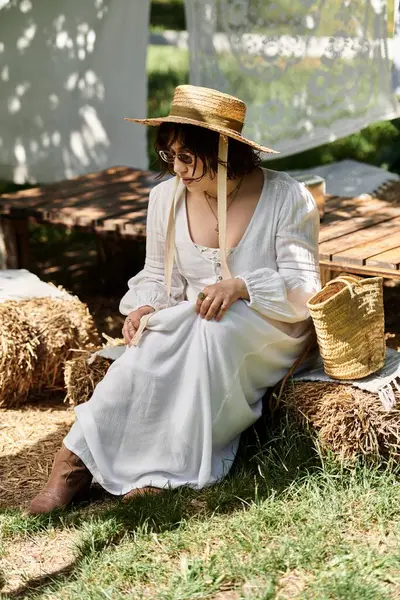 A young woman in a white dress and straw hat sits in a summer garden, enjoying the afternoon sun. — Stock Photo
