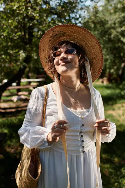 Una joven de cabello oscuro lleva una blusa blanca y un sombrero de paja, sonriendo mientras disfruta del sol de verano en un entorno de jardín.. - foto de stock