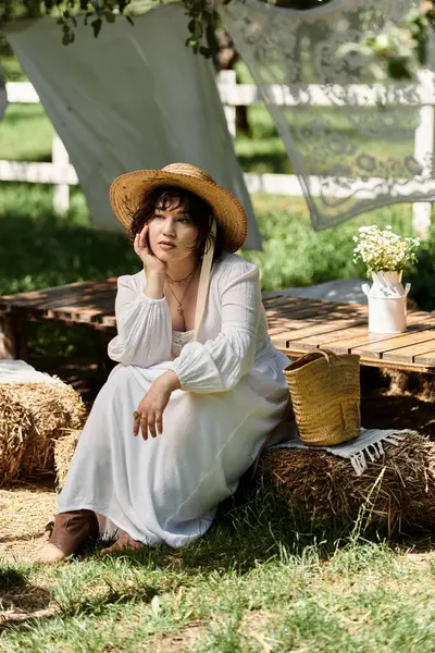 A young woman in a straw hat and white dress sits on a bale of hay in a summer garden. — Stock Photo