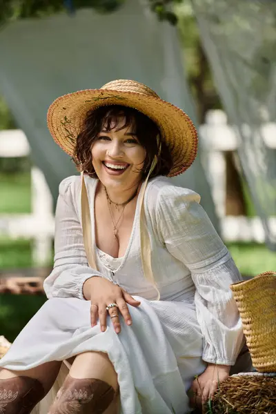 Uma jovem com cabelo escuro, vestindo um vestido branco e chapéu de palha, senta-se em um jardim, rindo. — Fotografia de Stock