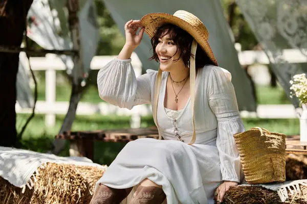 A young woman in a white dress and straw hat smiles brightly while sitting in a summer garden, enjoying the sunshine. — Stock Photo