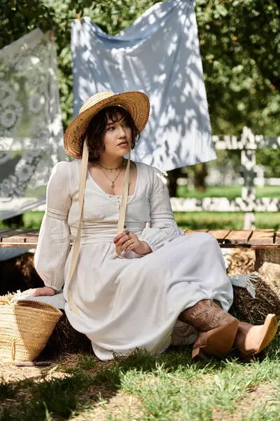 A young woman in a white dress and straw hat sits on a bale of hay in a summer garden. — Stock Photo