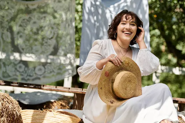 A young woman wearing a white dress and straw hat sits in a garden, smiling warmly as she holds her hat. — Stock Photo
