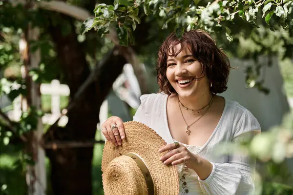A young woman with a straw hat and white blouse smiles brightly as she stands in a summer garden. — Stock Photo
