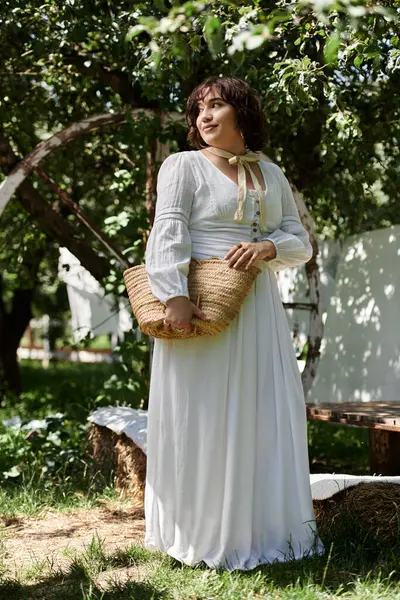 Una mujer joven con el pelo oscuro y un sombrero de paja se encuentra en un jardín de verano, con un vestido blanco y llevando una cesta tejida. - foto de stock