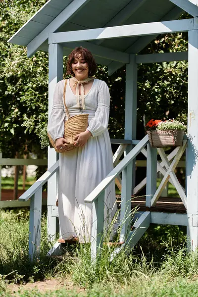 Uma jovem mulher em um vestido branco e chapéu de palha fica em um alpendre em um jardim exuberante. — Stock Photo