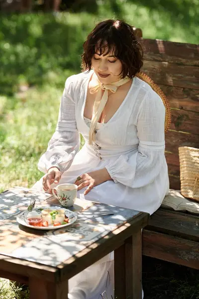 A young woman in a white dress enjoys a leisurely brunch in a summer garden, surrounded by lush greenery. — Stock Photo