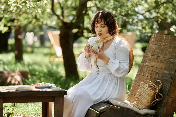 A woman in a white dress enjoys a cup of tea while sitting on a wooden bench in a summer garden. — Stock Photo