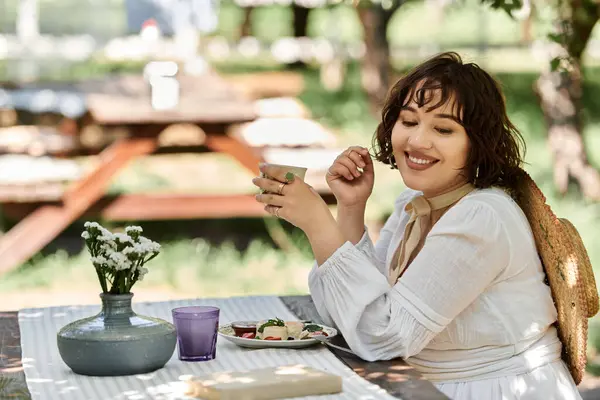 Uma bela morena desfruta de um brunch de lazer em um jardim ensolarado, vestido com um vestido branco. — Fotografia de Stock