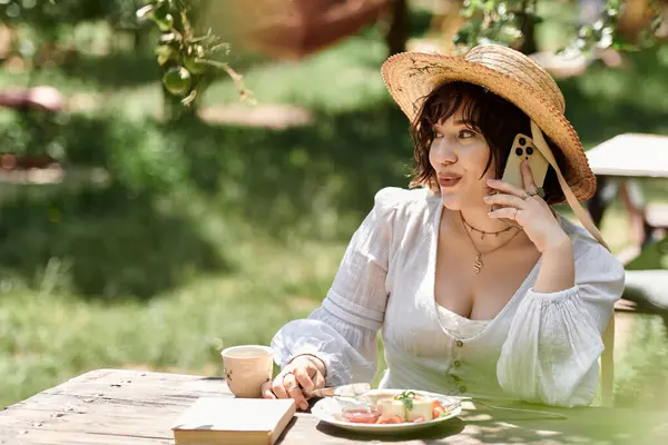 A young woman in a white dress enjoys a leisurely brunch outdoors, chatting on her phone while surrounded by lush greenery. — Stock Photo