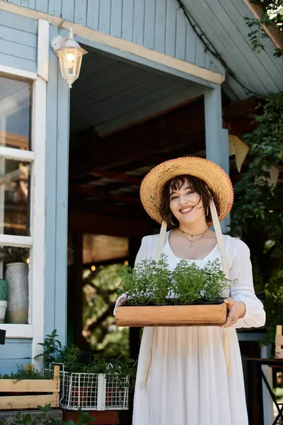 A young woman in a white dress and straw hat holds a tray of freshly potted plants, smiling at the camera. — Stock Photo