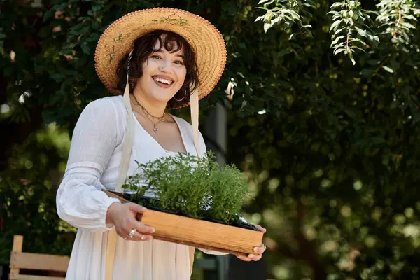 Una joven con un vestido blanco y sombrero de paja sonríe mientras lleva una bandeja de plantas en maceta en un exuberante jardín verde. — Stock Photo