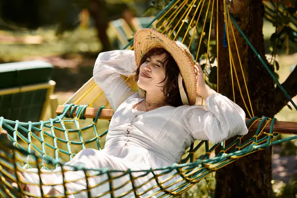 A young woman in a white dress and straw hat relaxes in a hammock under a summer sky. — Stock Photo