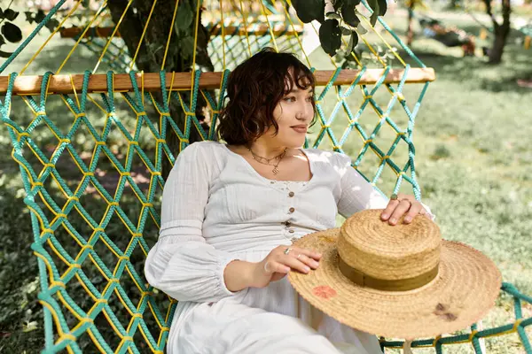 Uma jovem mulher em um vestido branco e chapéu de palha relaxa em uma rede sob a sombra de uma árvore. — Stock Photo