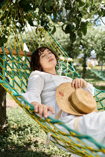 Uma jovem mulher em um vestido branco e chapéu de palha descansa pacificamente em uma rede, cercada por uma exuberante folhagem verde.. — Fotografia de Stock